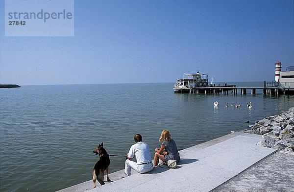 Paar mit Hund an der K??ste mit Pier im Hintergrund  Neusiedler See Lake  Podersdorf  Burgenland  Österreich