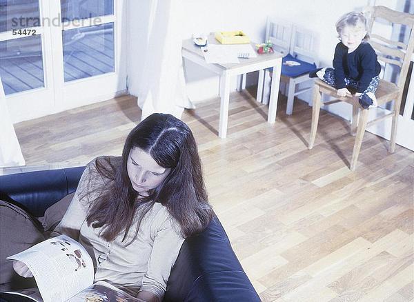 Erhöhte Ansicht von Frau Lesung Buch über Sofa mit ihrem Kind sitzt auf Stuhl im Hintergrund