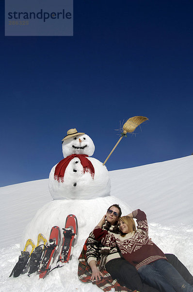 Paar sitzend bei Schneemann mit Schneeschuhen