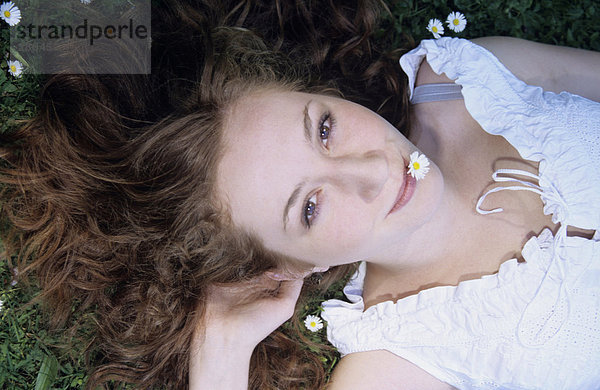 Junge Frau mit Gänseblümchen im Mund auf Gras liegend  Nahaufnahme  Portrait