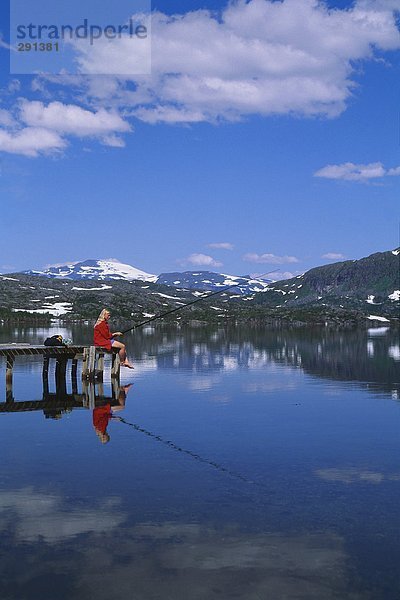 Eine Frau Angeln von einer Brücke durch einen ruhigen See in den Bergen.