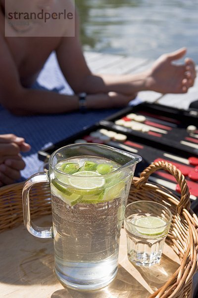 Eine Kanne Wasser und ein Glas mit Stücken von Kalk auf einem Tablett ein Spiel von Backgammon im Hintergrund.