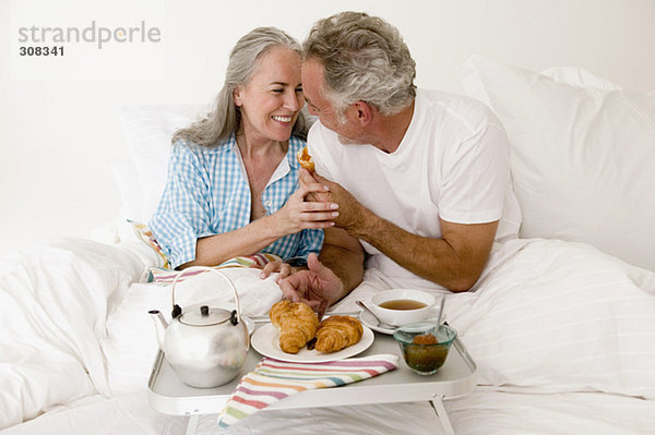 Erwachsenes Paar auf dem Bett sitzend mit Frühstück  lächelnd