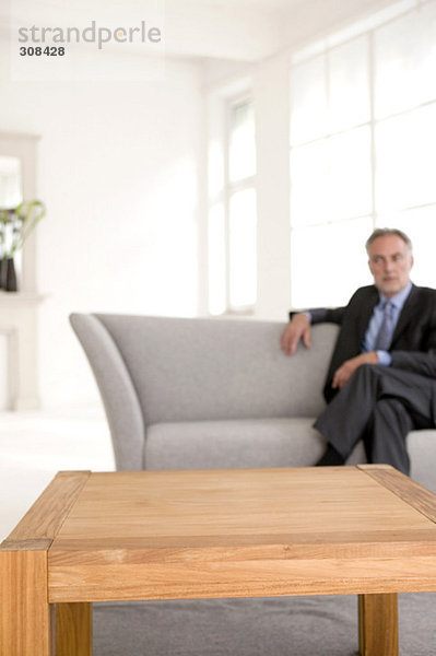 Erwachsener Mann sitzt auf dem Sofa und schaut weg (Fokus auf den Tisch im Vordergrund)