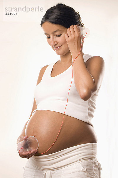 Schwangere Frau hält Tasse an Bauch und Ohr