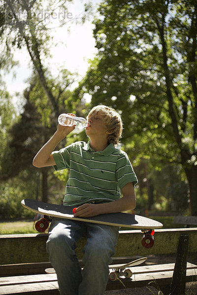 Junge Trinken von eine Flasche mit einem Skateboard im Knie.