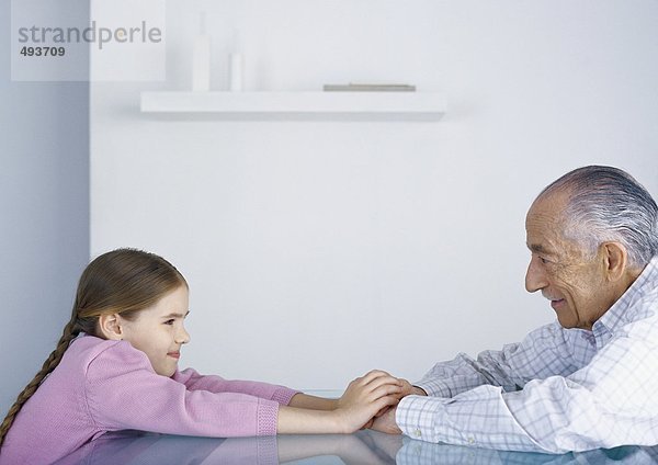 Mädchen und Großvater sitzen sich am Tisch gegenüber und halten Händchen.
