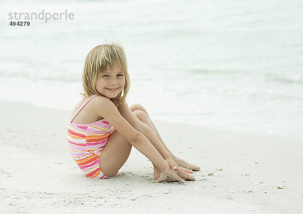 Kleines Mädchen sitzt am Strand mit erhobenen Knien und lächelt in die Kamera.