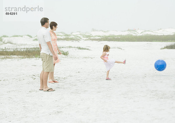 Familie am Strand  Eltern sehen zu  wie ein kleines Mädchen den Ball kickt.