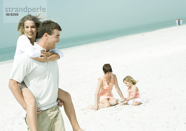 Familie am Strand  Mann mit Teenager-Tochter Huckepack  während Mutter mit Tochter auf Sand sitzt.