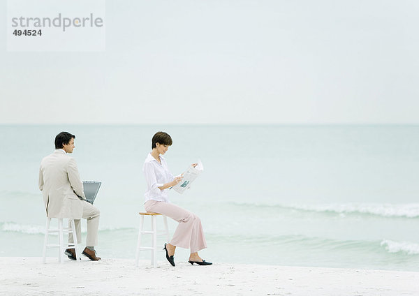 Geschäftsleute sitzen auf Hockern am Strand  einer mit Laptop  ein anderer mit Zeitung lesen