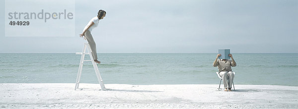 Am Strand  Frau steht auf einer Leiter und schreit den Mann an  der sitzt und das Gesicht bedeckt.