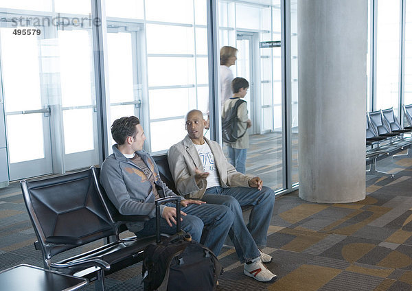 Zwei Männer sitzen in der Flughafenlounge  Reisende gehen im Hintergrund vorbei.