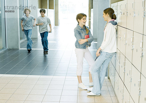 Zwei Teenagermädchen reden an Spinden  während Jungen durch den Flur rennen.