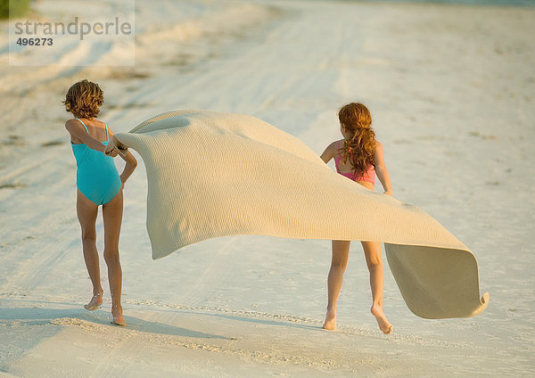 Zwei Mädchen laufen am Strand und halten die Decke im Wind.
