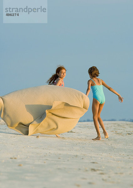 Zwei Mädchen laufen am Strand und halten die Decke im Wind.