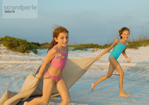 Zwei Mädchen laufen am Strand und halten die Decke im Wind.
