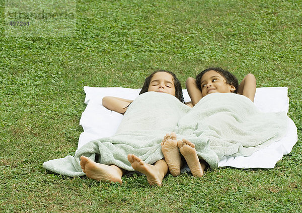 Zwei Mädchen liegen im Gras  auf einem Handtuch und mit einem Handtuch bedeckt.