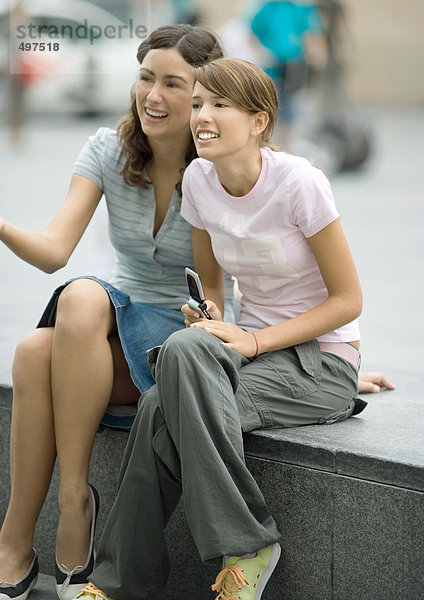Zwei Teenager-Mädchen sitzen auf einer Bank in städtischer Umgebung  lachend  eine hält ein Handy in der Hand