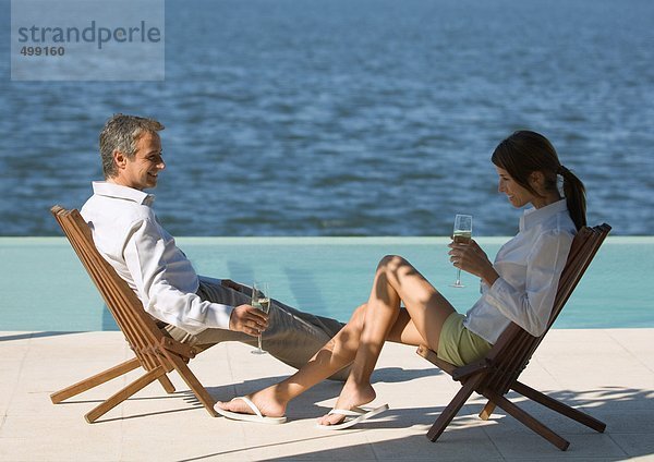 Paar sitzt in Liegestühlen am Meer und trinkt Champagner.