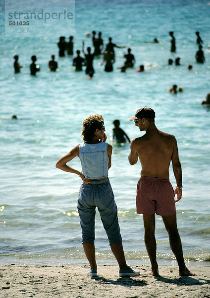 Leute stehen am Strand  Leute spielen im Wasser im Hintergrund  Rückansicht.