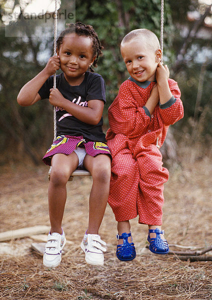Kleiner Junge und Mädchen sitzen auf einer Schaukel  jede hält eine Seite des Schaukelseils  lächelnd vor der Kamera  volle Länge.