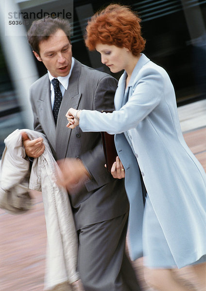Geschäftsmann und Geschäftsfrau gehen Seite an Seite nach draußen  Geschäftsfrau schaut auf die Uhr