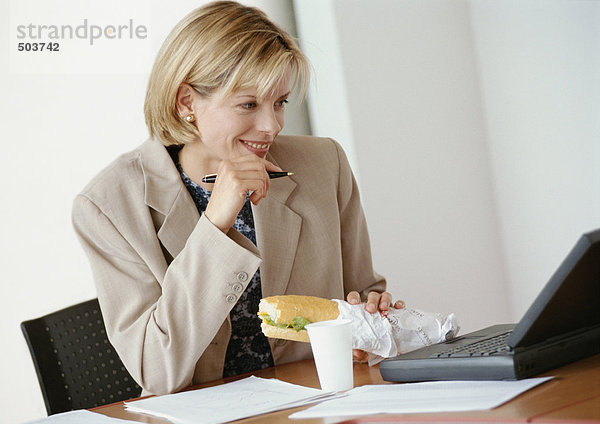 Geschäftsfrau am Schreibtisch sitzend mit Laptop-Computer  Sandwich haltend