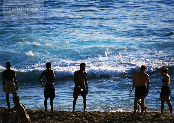 Silhouetten von Menschen  die am Strand am Rande des Meeres stehen  Wellen im Hintergrund