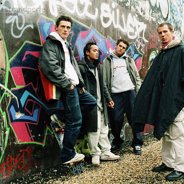 Vier junge Männer in der Stadtstraße  drei lehnen sich an die Graffiti-Mauer.