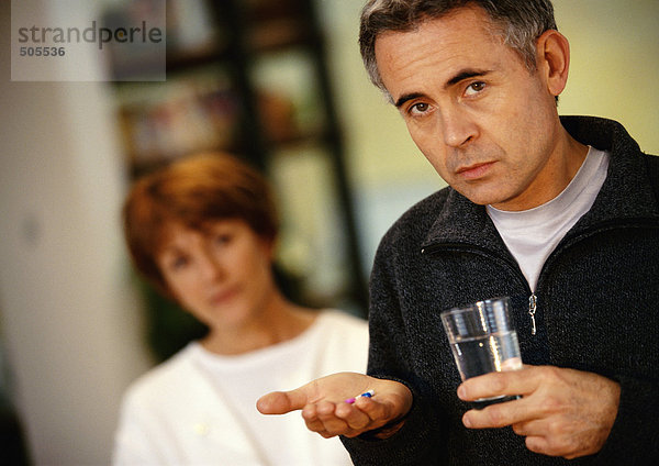 Mann hält Glas in der einen Hand und Pillen in der anderen  Frau verschwommen im Hintergrund