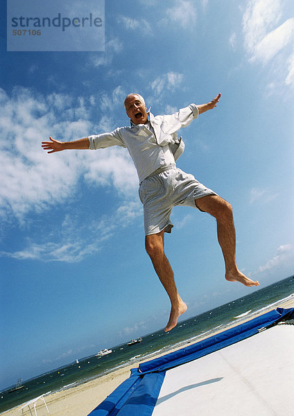 Erwachsener Mann springt auf Trampolin am Strand  Arme ausstrecken  Mund offen