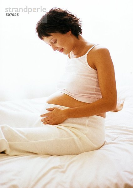 Eine schwangere Frau sitzt auf dem Bett und schaut auf ihren Bauch.