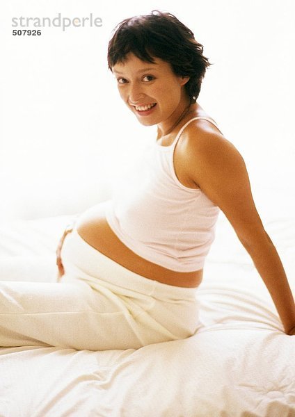 Schwangere Frau auf dem Bett sitzend  lächelnd vor der Kamera  Porträt
