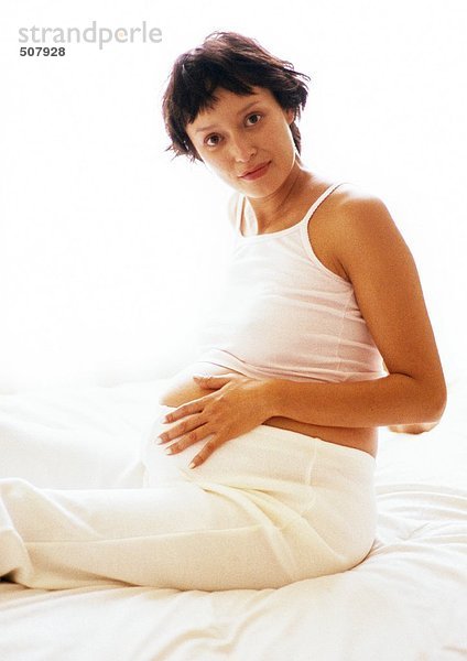 Schwangere Frau sitzt mit der Hand auf dem Bauch  schaut in die Kamera  Porträt