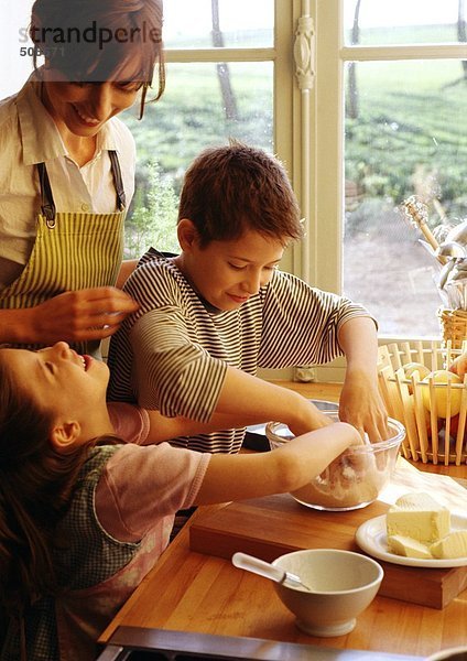 Zwei Kinder und Mutter kochen in der Küche  Kinder mit Händen in der Schüssel  Mutter in der Schürze lächelnd