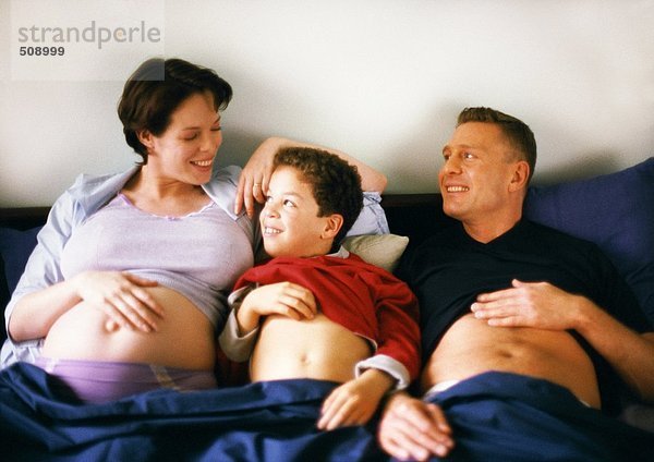 Schwangere Frau mit Mann und Kind im Bett  die alle ihre Bäuche zeigen.