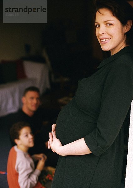 Schwangere Frau stehend und haltend  Mann und Kind sitzend im Hintergrund