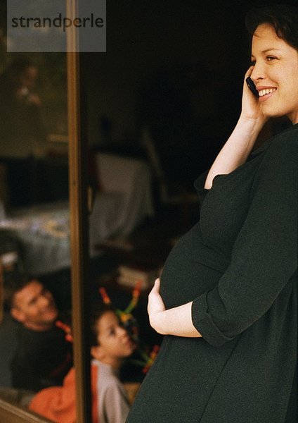 Schwangere Frau im Stehen  mit dem Handy und mit dem Bauch  Mann und Kind im Hintergrund sitzend.