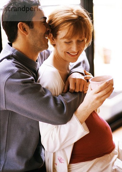 Mann umarmt schwangere Frau von hinten  lächelnd