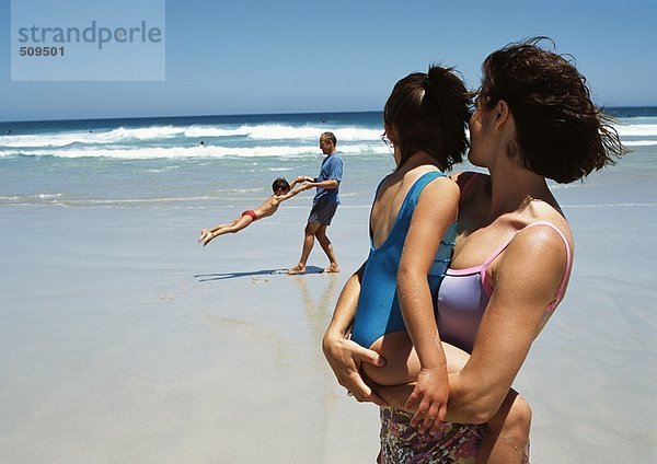 Frau hält kleines Mädchen in den Armen im Vordergrund am Strand  Mann schwingt kleinen Jungen im Hintergrund