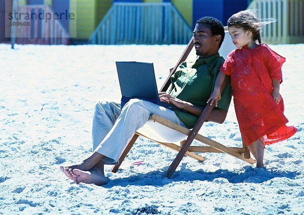 Mann sitzt auf einem Stuhl am Strand mit Laptop  kleines Mädchen steht an seiner Seite