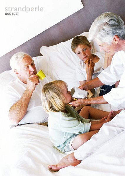 Großeltern und Kinder beim Frühstücken im Bett  Frau hilft dem kleinen Mädchen beim Trinken aus der Tasse