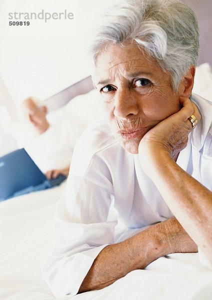 Seniorin im Bett mit Hand unter dem Kinn  Mann mit Laptop im Hintergrund