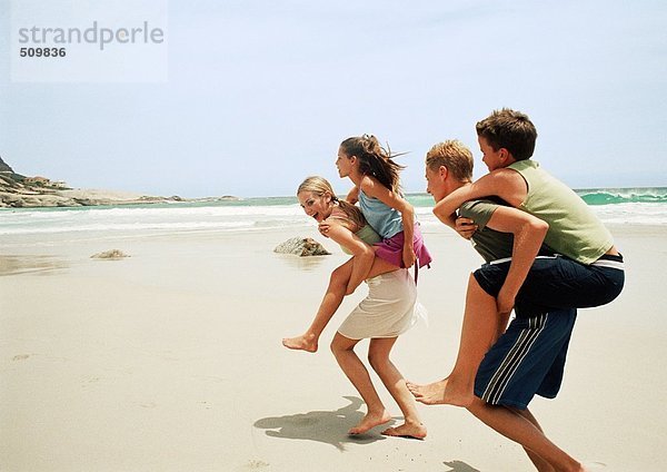 Zwei junge Leute  die Kinder huckepack am Strand tragen.