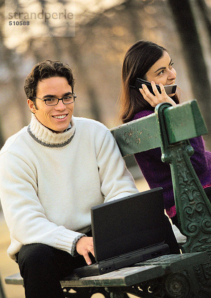 Mann sitzt auf Bank mit Laptop und Frau mit Handy