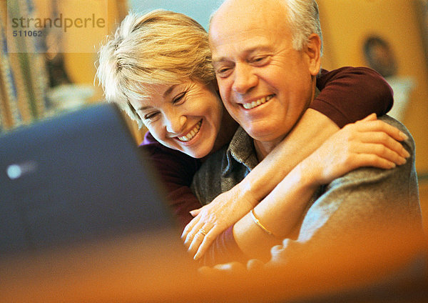 Älteres Paar lächelnd  Mann mit Laptop  Frau mit Armen um den Mann herum