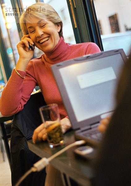 Reife Frau  die ein Handy benutzt  sitzt am Tisch gegenüber von jemandem  der einen Laptop benutzt.