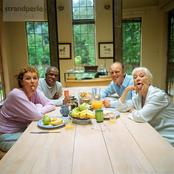 Vier reife Menschen  die am Tisch sitzen und die Zungen herausstrecken.