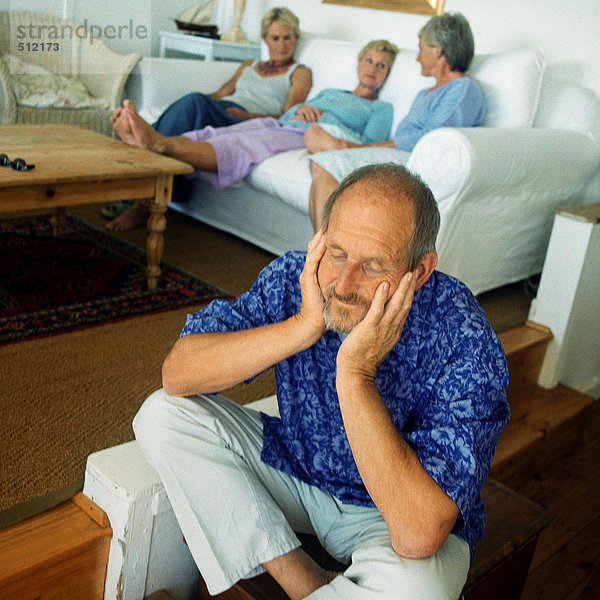 Erwachsener Mann sitzt auf dem Boden  drei Frauen im Hintergrund auf der Couch.